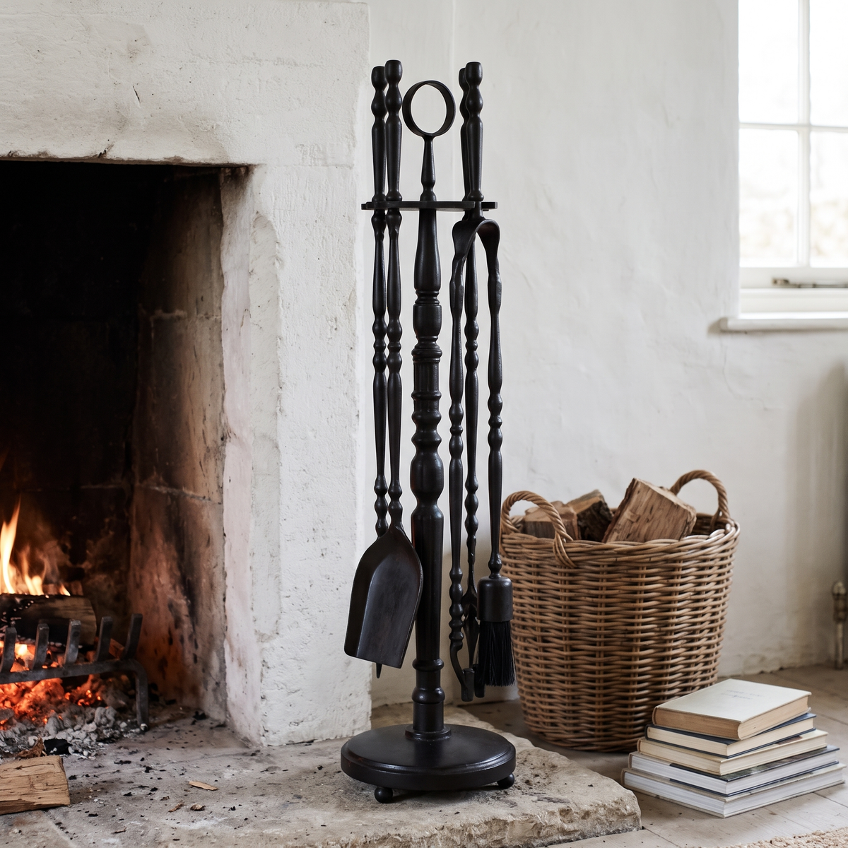 Black brass fireplace tools beside a limestone fireplace with a fire and a log basket