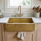 Brass farmhouse apron sink below a window in a bright white oak kitchen