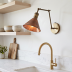 Brass swing arm sconce with copper shade over a marble kitchen counter beside open shelves