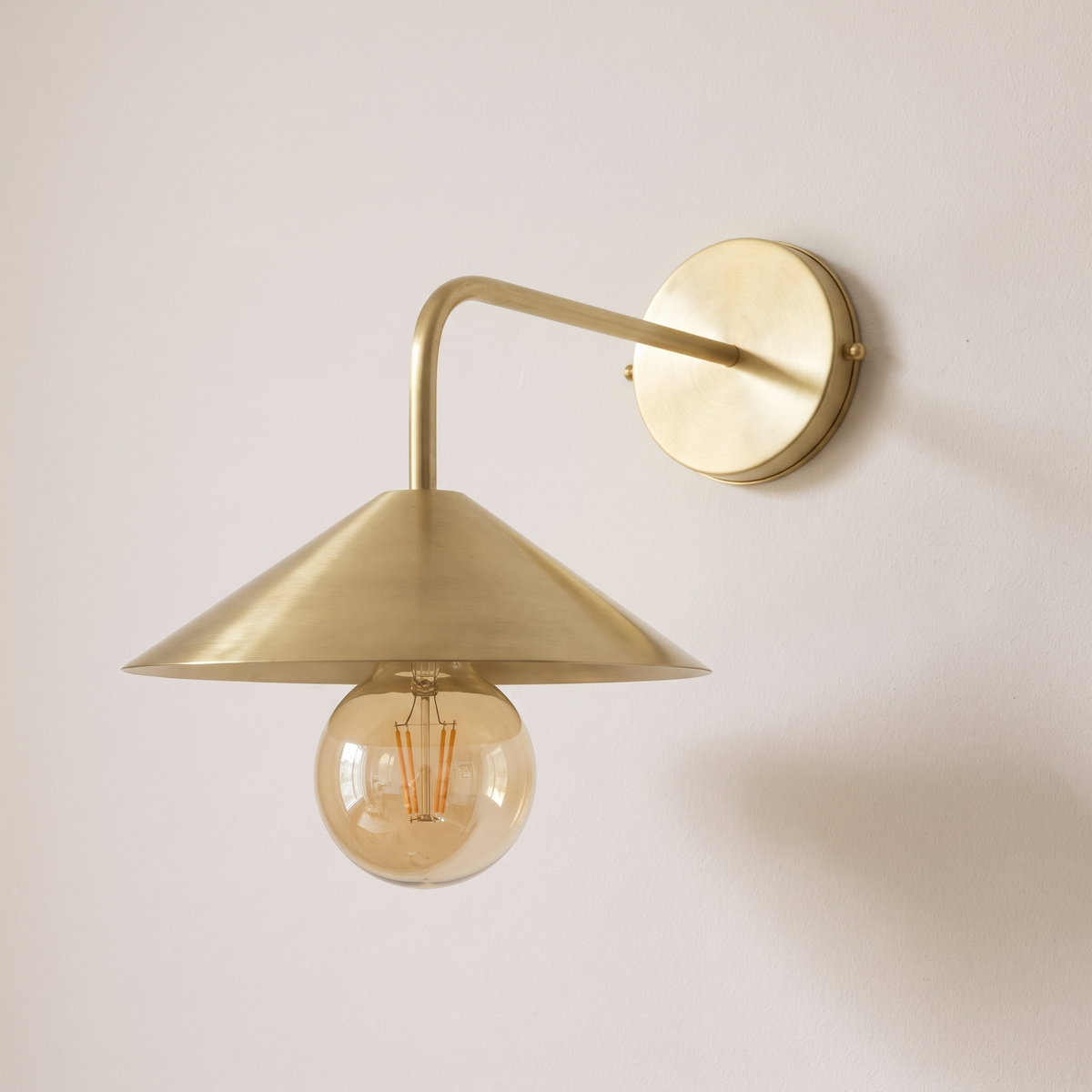 Brushed brass angled arm wall sconce with cone shade and globe bulb on a light background