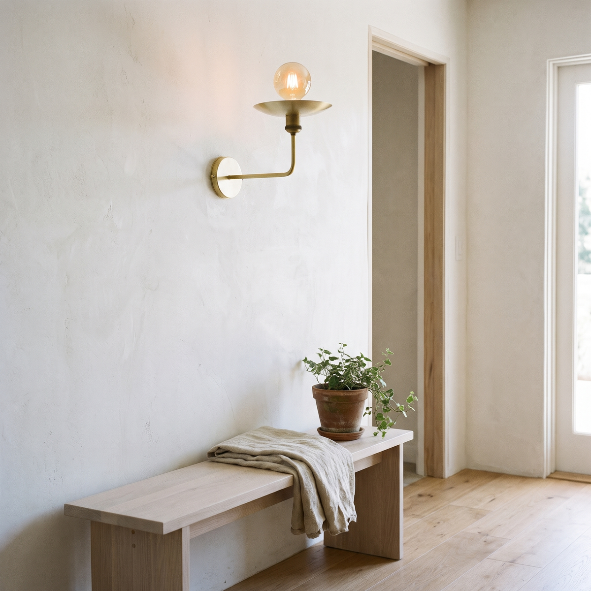 Brushed brass uplight saucer sconce on a plaster wall above a white oak bench