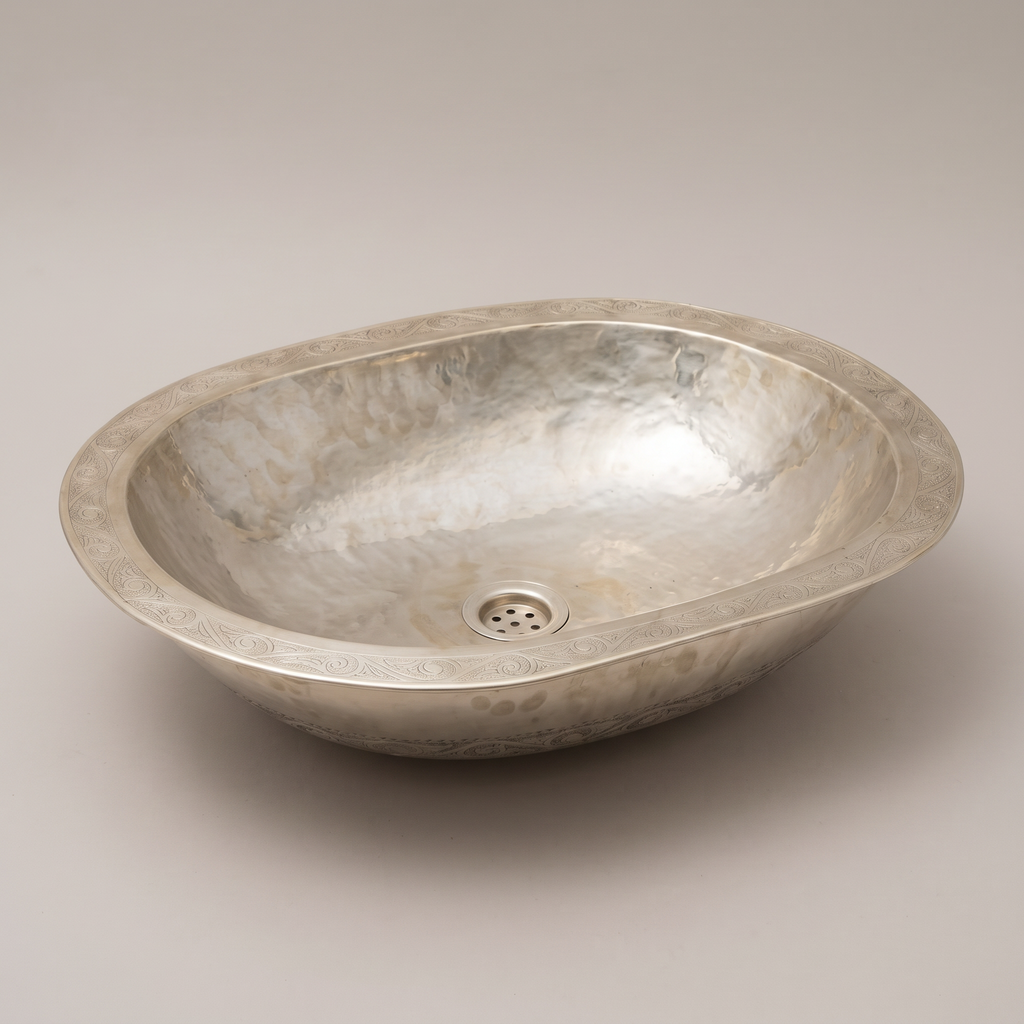Low oval German silver vessel sink with engraved rim and hammered basin on a plain background