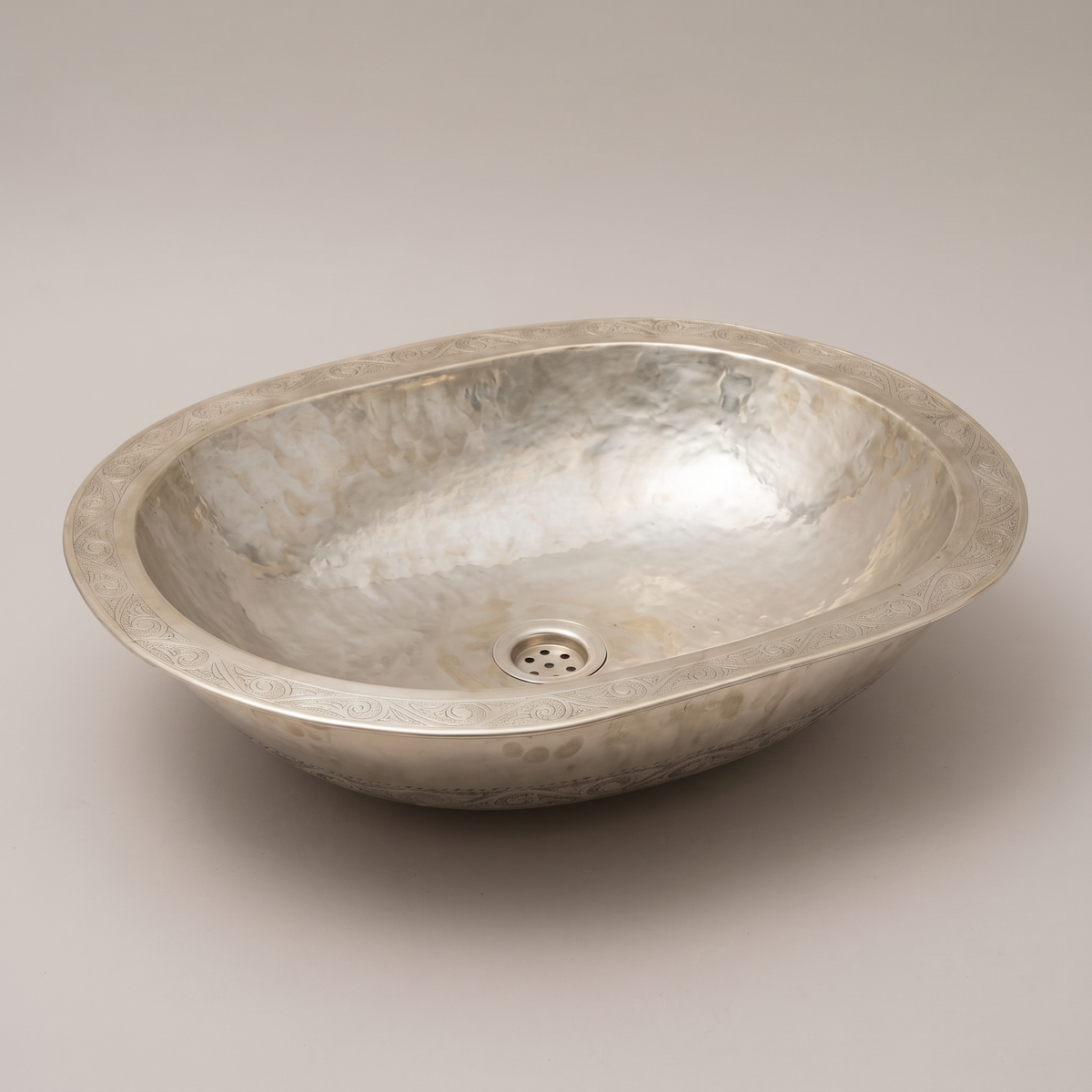 Low oval German silver vessel sink with engraved rim and hammered basin on a plain background