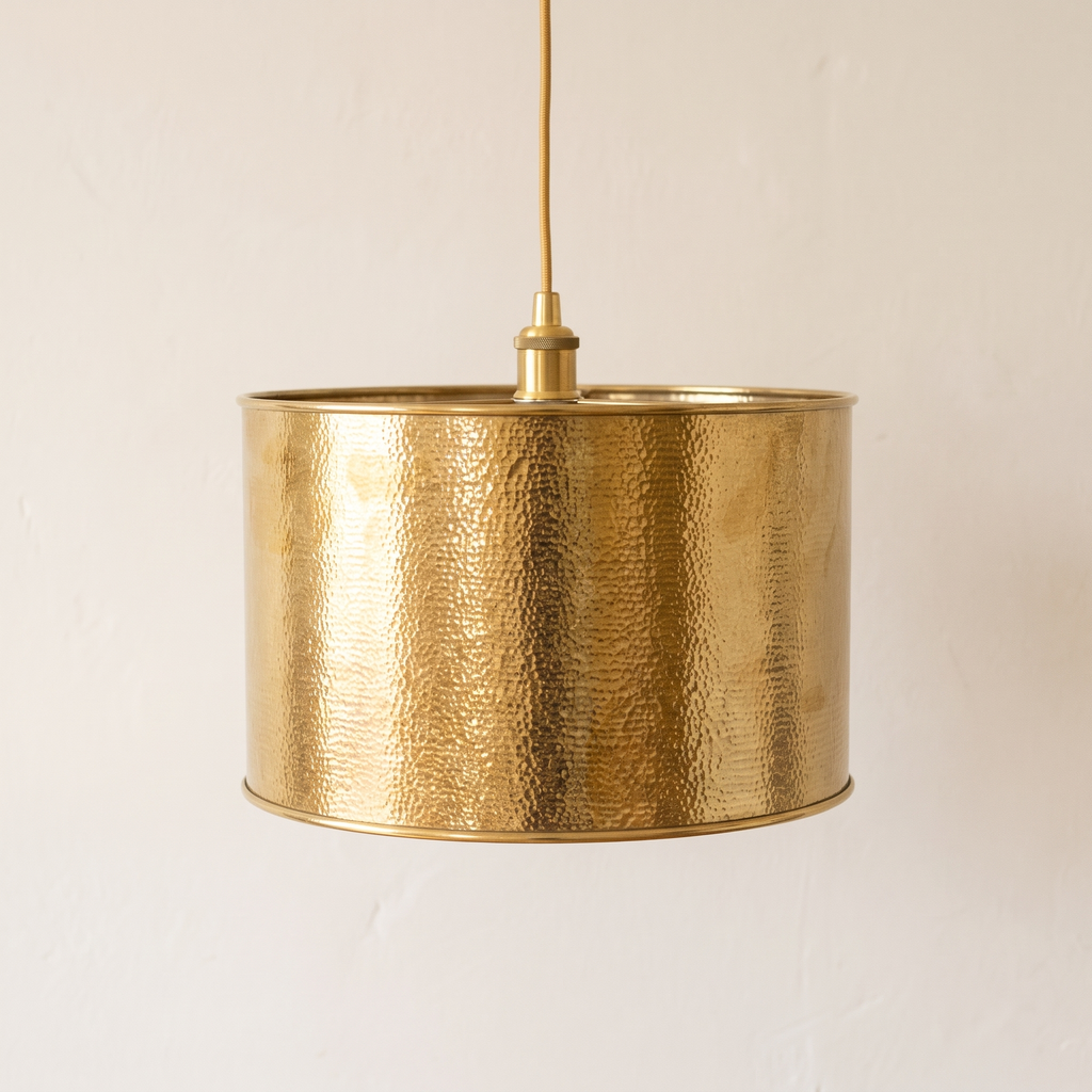 Hammered unlacquered brass drum pendant with gold cord on a light background