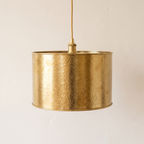 Hammered unlacquered brass drum pendant with gold cord on a light background