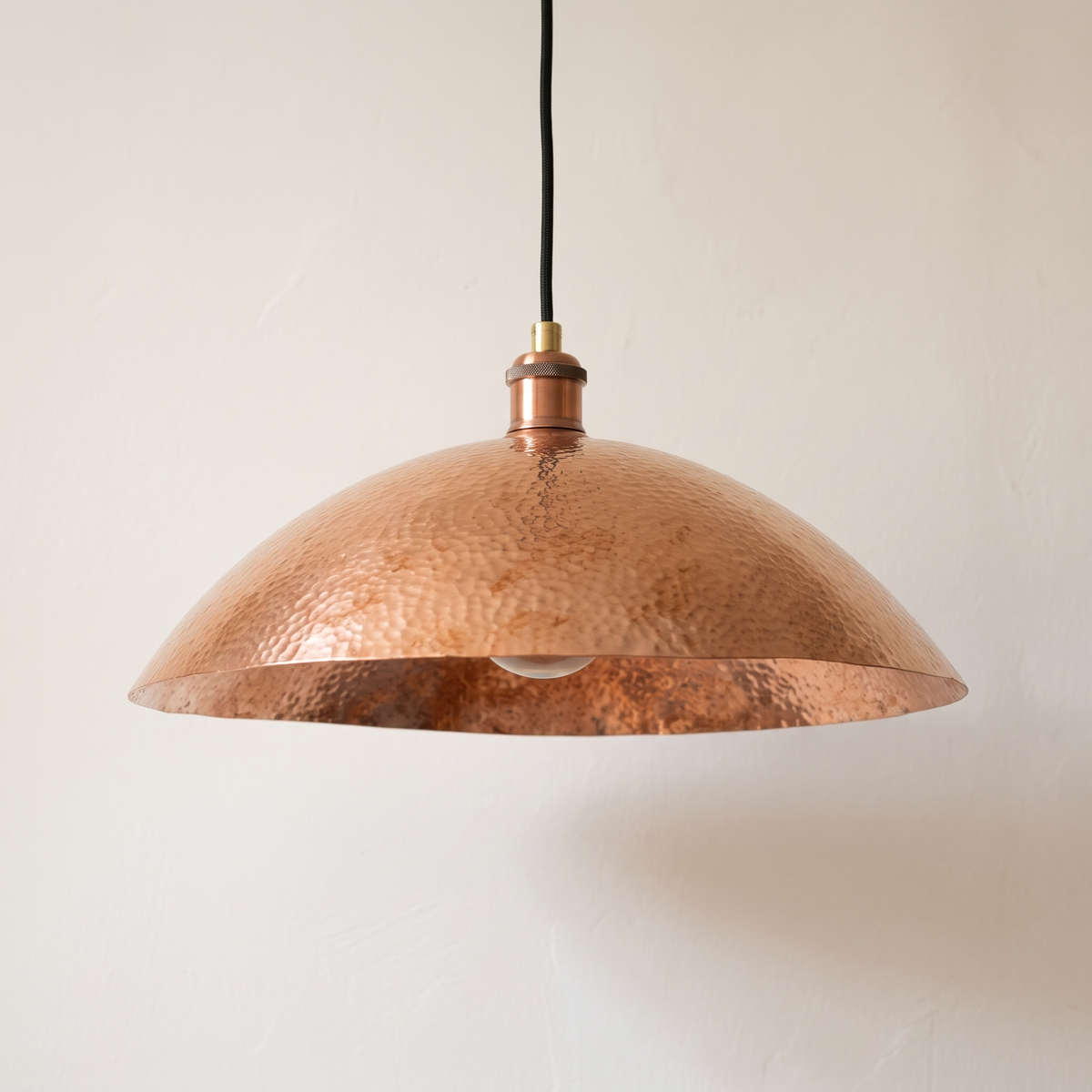 Hammered unlacquered copper wide dome pendant with black cord on a light background