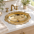 Hammered oval brass drop-in sink with scalloped rim in a sunlit white marble vanity
