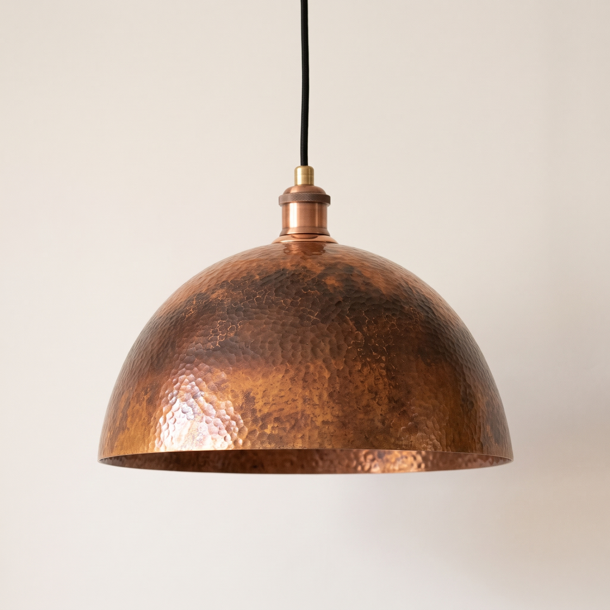 Hammered copper dome pendant with oxidized patina and black cord on a light background