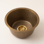 Round aged brass prep sink with tapered sides and a brass basket strainer, on a light background