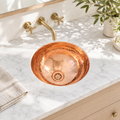 Hammered round copper undermount sink in a sunlit white marble bathroom vanity