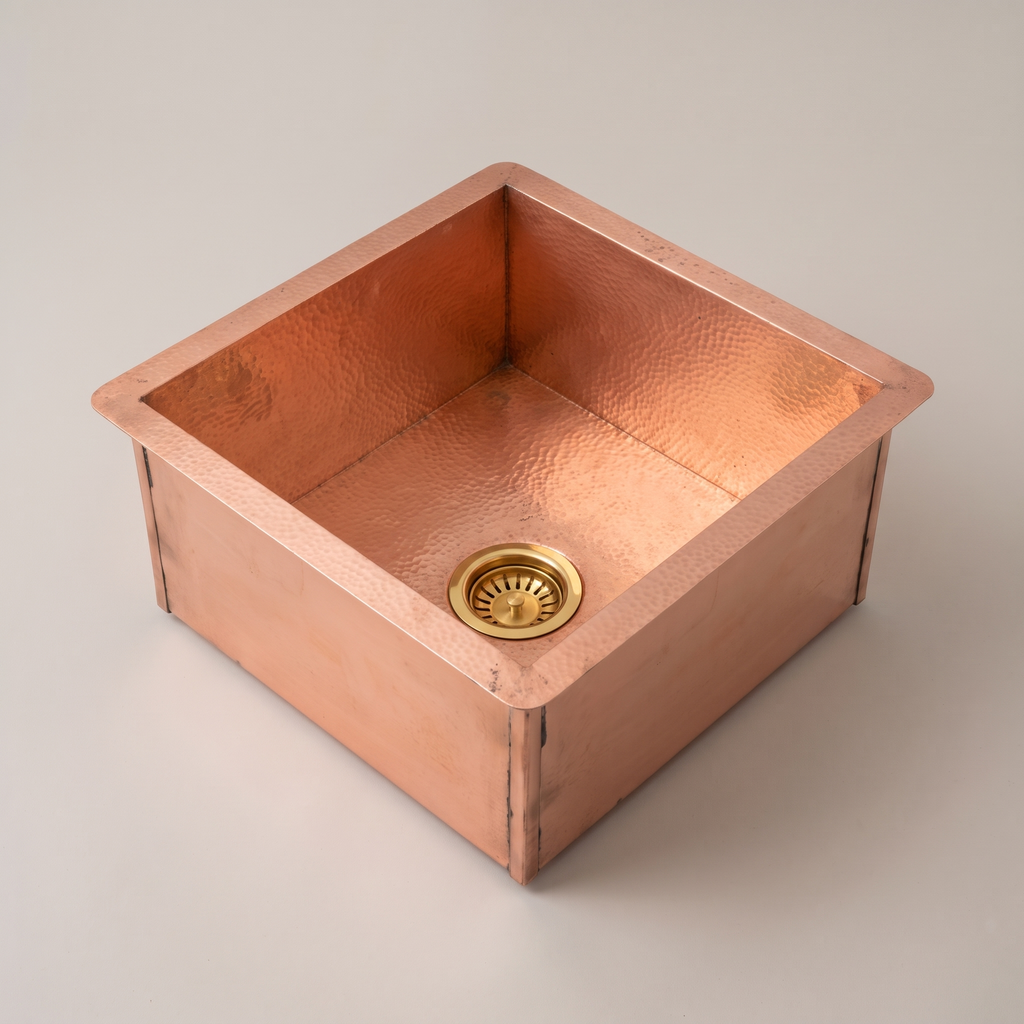 Square copper undermount sink with hammered flange, visible seams and brass basket strainer on a plain background